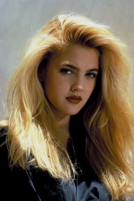 A young woman with long, voluminous blonde hair and dark lipstick looks seriously at the camera. She is wearing a dark top and is posed against a light, softly textured background.