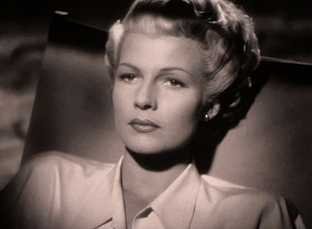 A black-and-white close-up of a woman with light hair styled in soft waves, wearing a collared blouse, gazing thoughtfully slightly off-camera with a serious expression.