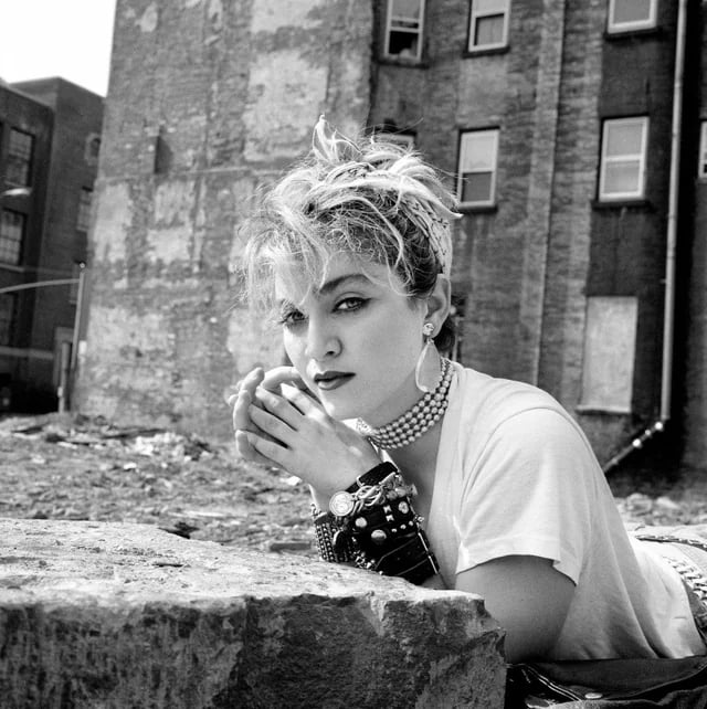 A woman with short, tousled blonde hair and bold makeup leans on a stone in an urban setting, wearing a white T-shirt, chunky jewelry, and layered bracelets. Worn brick buildings stand in the background.