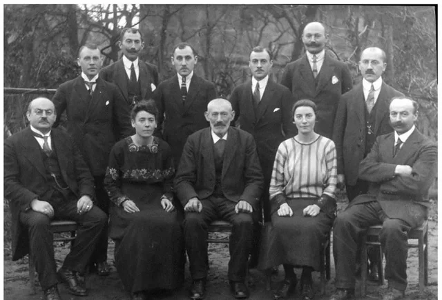 A black and white photo of eleven people from the early 20th century, including nine men in suits and two women in long skirts and blouses, seated and standing in two rows outdoors.