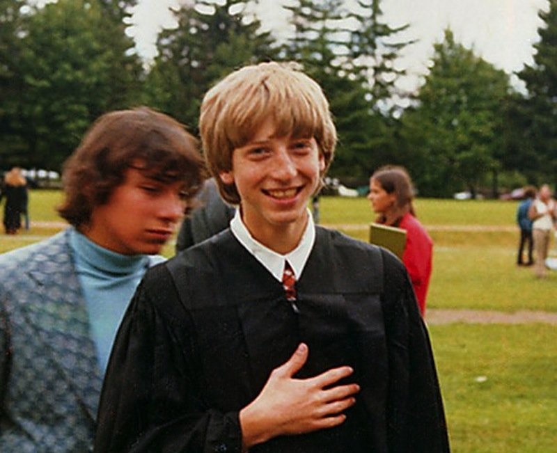 A young person in a black graduation gown smiles at the camera outdoors, with trees and other people in the background. Another person in a blue shirt and vest walks by, slightly blurred.