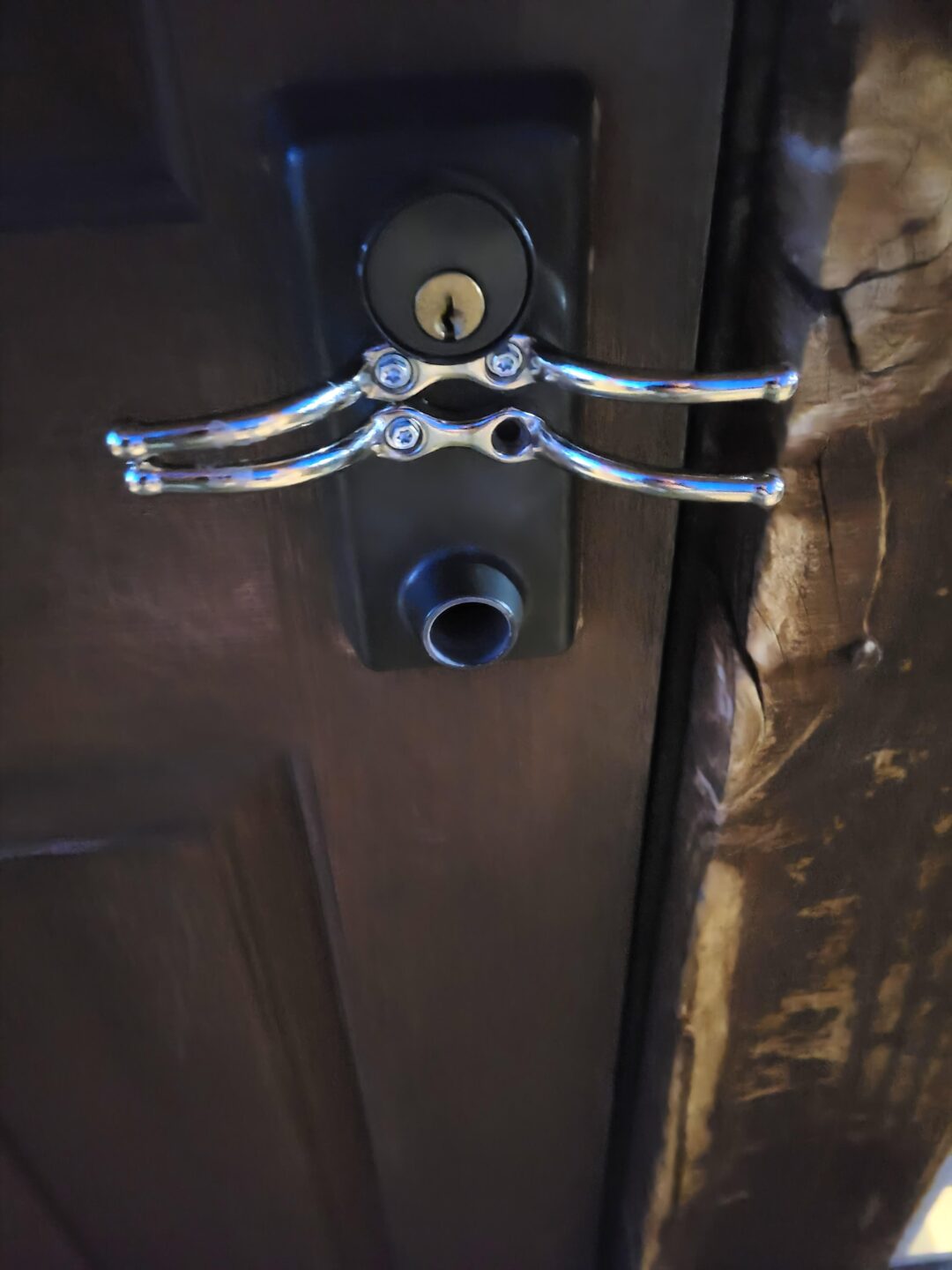 A close-up of a dark wooden door with a unique metallic door handle that resembles a pair of pliers, mounted above a circular keyhole and a cylindrical door lock. The wood around the lock appears worn.