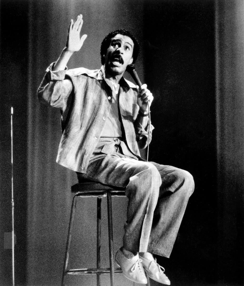 A man sits on a stool on stage, holding a microphone in one hand and raising his other hand, as if making a point while performing. He wears a casual suit and sneakers. The image is in black and white.