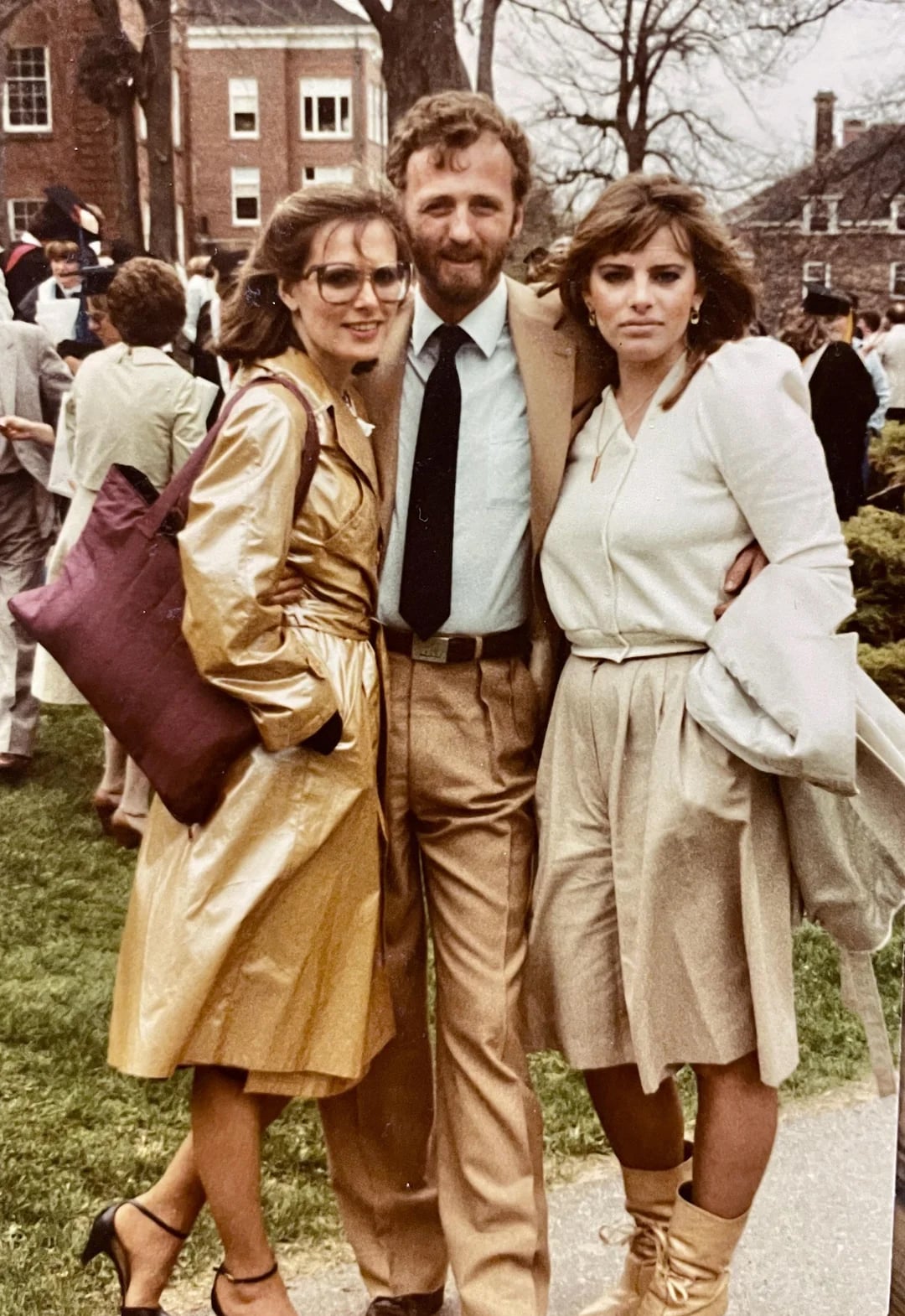 Three adults dressed in tan and cream 1980s-style outfits pose together outdoors at a formal event, with buildings, other people, and trees visible in the background.