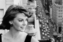 A woman in a black dress stands on a balcony overlooking a busy city street lined with tall buildings. She smiles while the wind blows her hair, and cars fill the street below. The scene appears to be from a bygone era.
