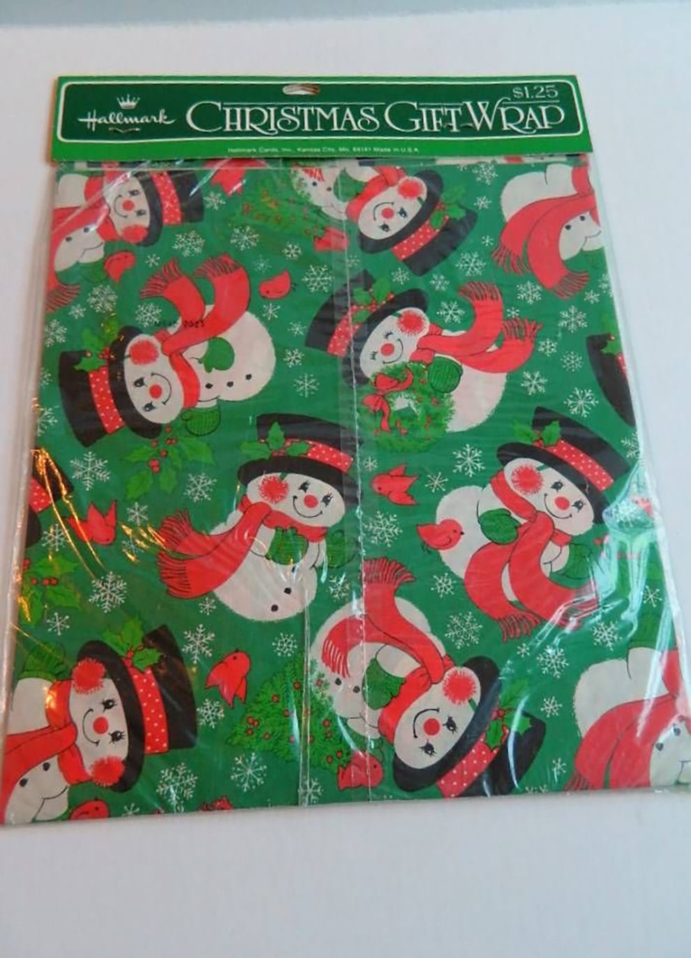 A package of Hallmark Christmas gift wrap featuring a festive pattern of snowmen wearing red scarves and black hats on a green background with snowflakes and red birds. The price tag shows $1.25.