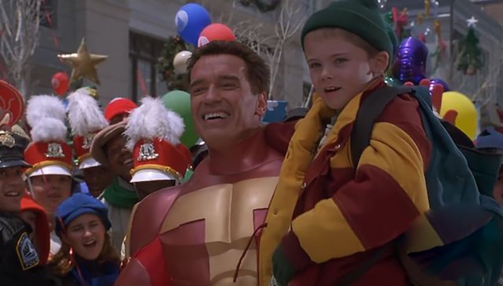 A man in a superhero costume smiles while carrying a young boy in colorful winter clothes. They are surrounded by a festive crowd with balloons and holiday decorations in the background.