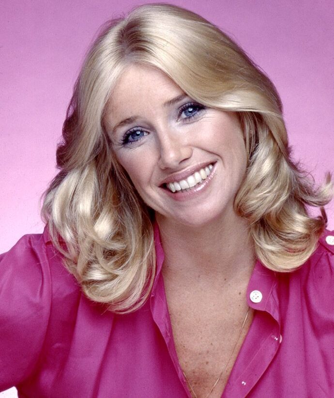 A smiling woman with blonde, shoulder-length hair wearing a bright pink blouse poses in front of a pink background.