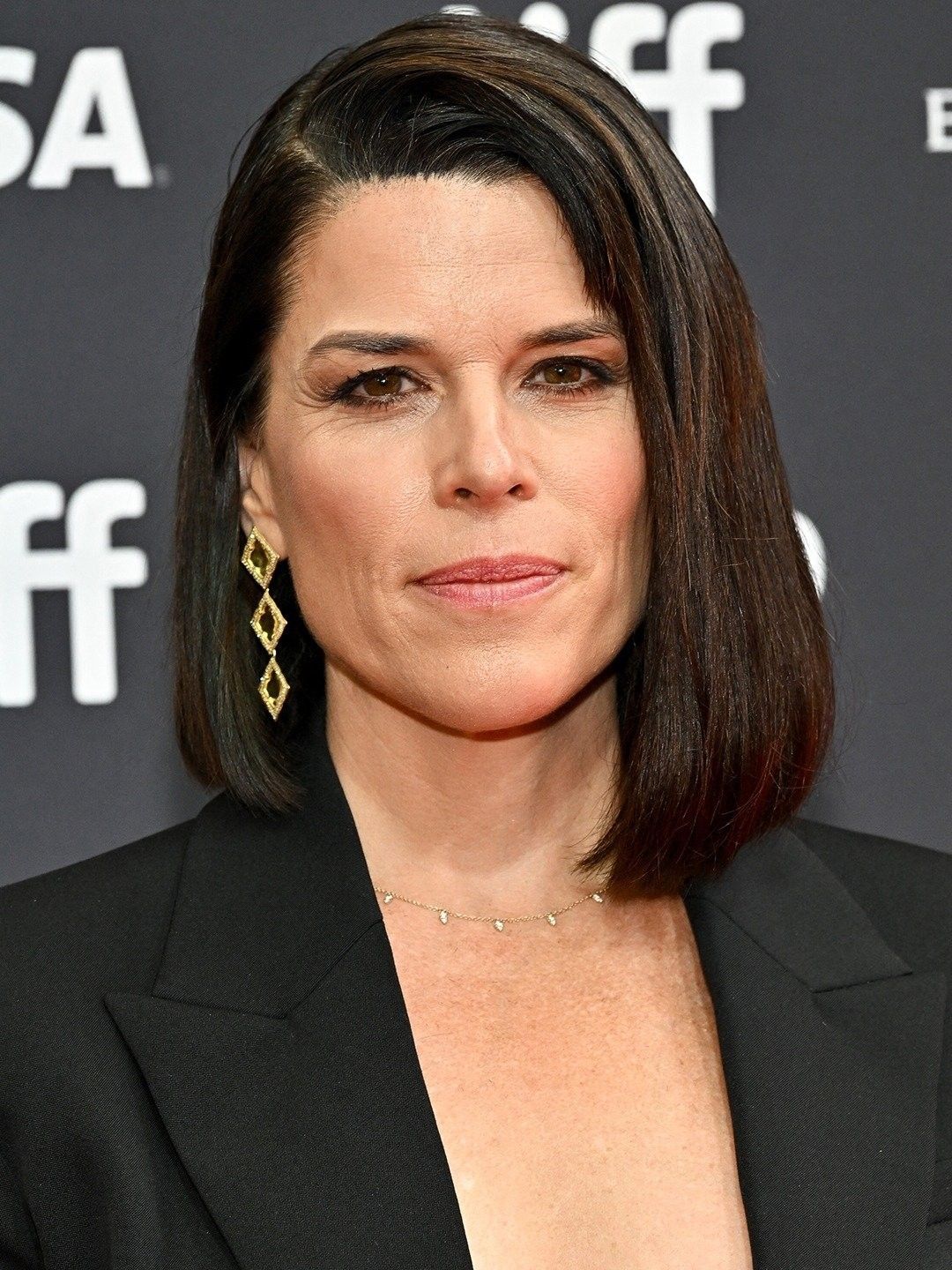 A woman with straight, dark shoulder-length hair wearing a black blazer, gold geometric earrings, and a delicate necklace poses in front of a black event backdrop with white text.