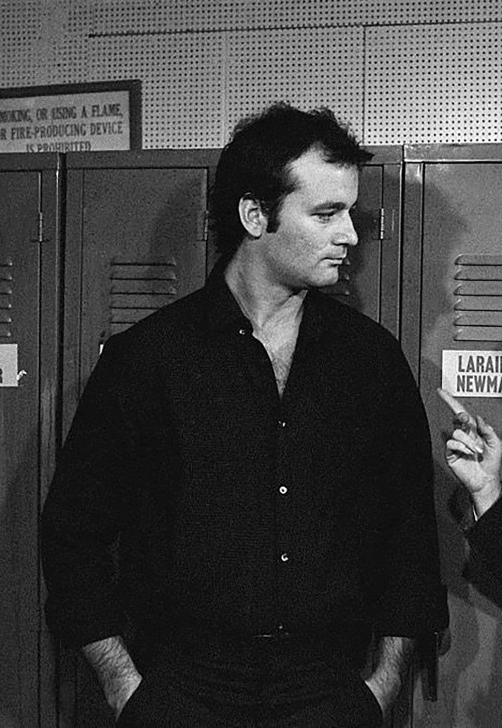 A man with dark hair, wearing a black shirt, stands in front of a row of lockers, looking to his right. A partially visible hand gestures beside him. A sign on a locker reads "LARAINE NEWM.