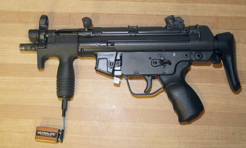 A compact black submachine gun with a vertical foregrip, folding stock, and an attached Ultralife battery pack, displayed on a wooden surface.