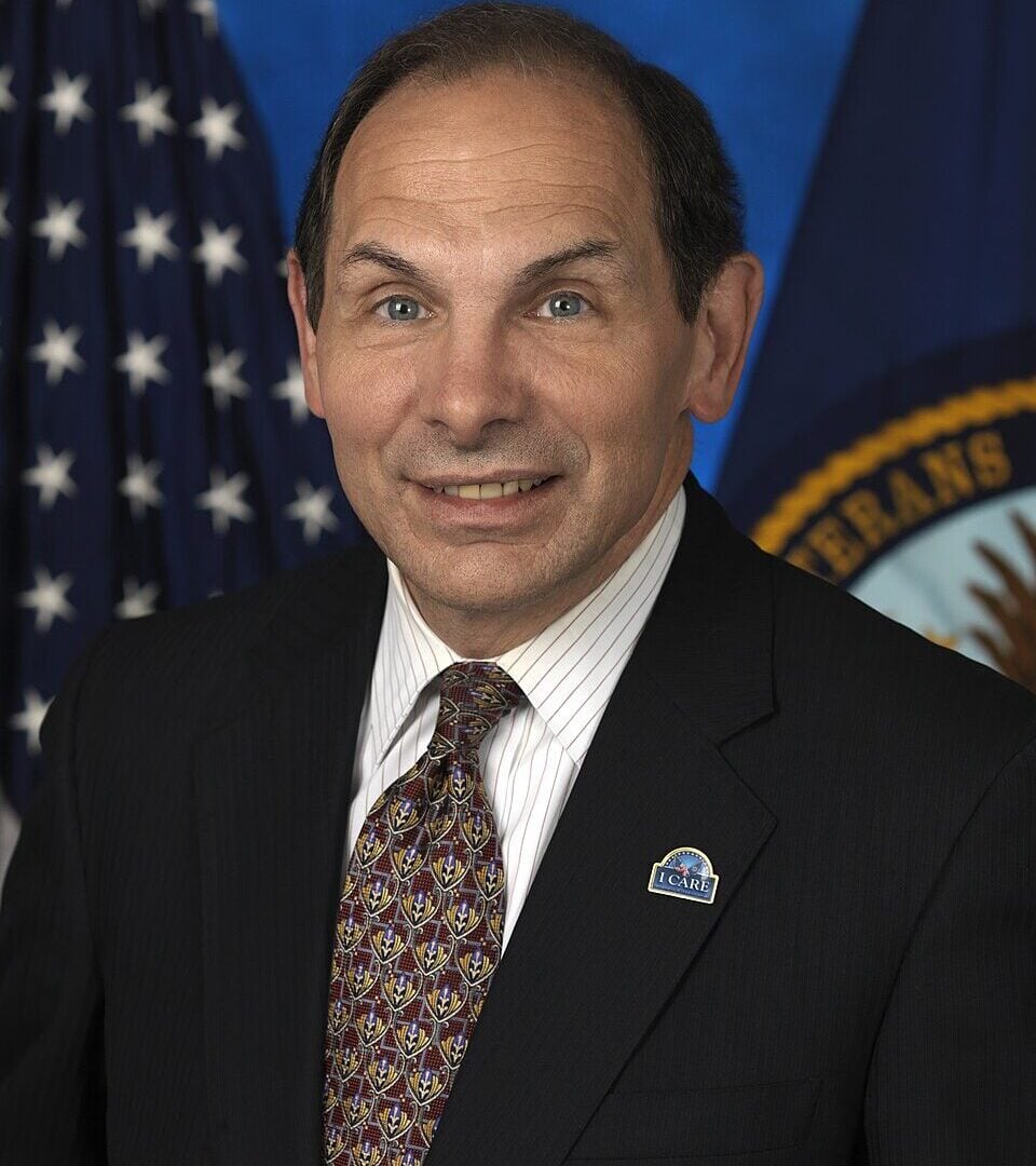A man in a dark suit, patterned tie, and striped shirt smiles in front of an American flag and a blue flag with a seal. He has short dark hair and is wearing a pin on his lapel.