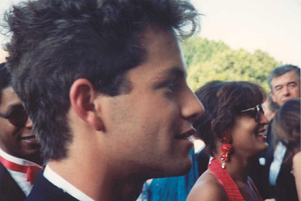 A young man in a suit is shown in profile at an outdoor event, with people dressed formally in the blurred background, including a woman in red earrings and sunglasses. Trees can be seen in the distance.
