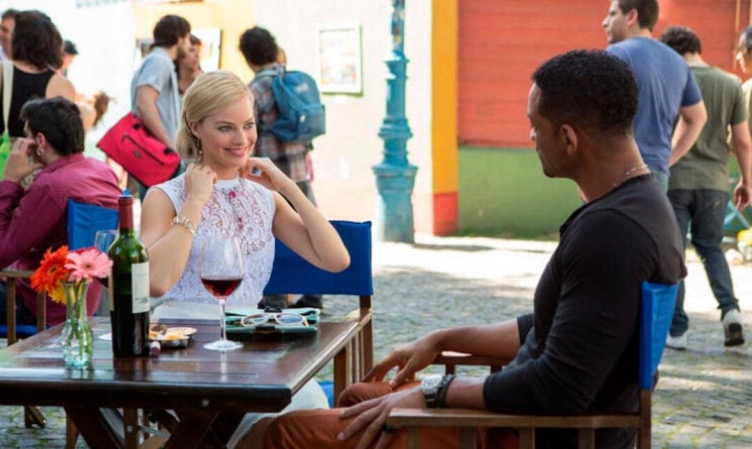 A woman in a white lace top and a man in a black shirt sit at an outdoor café table with wine, talking and smiling, while people walk and socialize in the background on a sunny day.