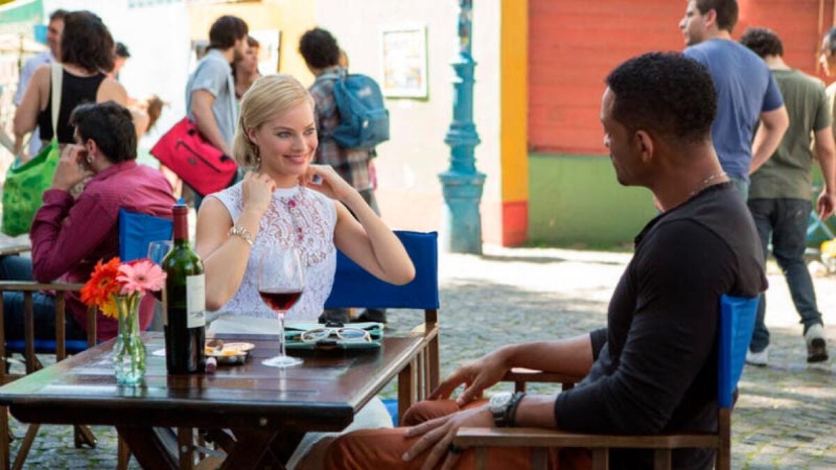 A woman in a white dress and a man in a dark shirt sit at an outdoor café table with wine, talking and smiling. People walk in the background on a sunny day.