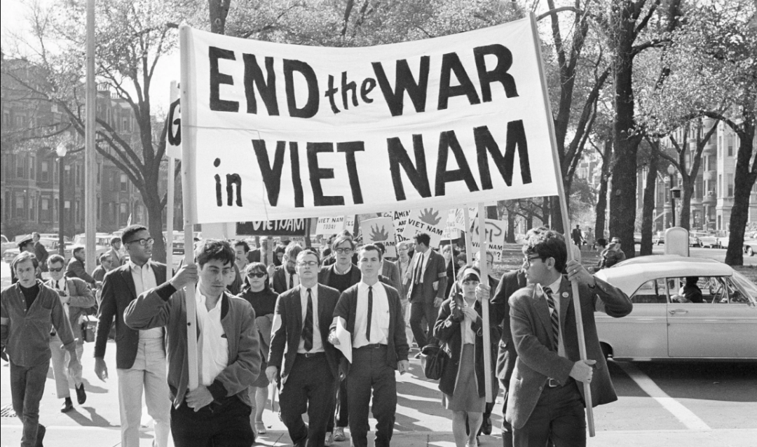 A group of protesters march down a city street holding a large banner that reads "END the WAR in VIET NAM." They are dressed in 1960s attire, and cars and trees are visible in the background.