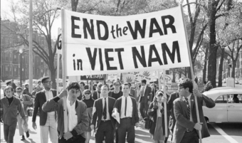 A group of protesters march down a city street holding a large banner that reads "END the WAR in VIET NAM." They are dressed in 1960s attire, and cars and trees are visible in the background.
