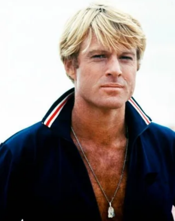 A blonde man with a serious expression wears a navy shirt with a red, white, and blue striped collar and a necklace, standing against a pale, blurred background.