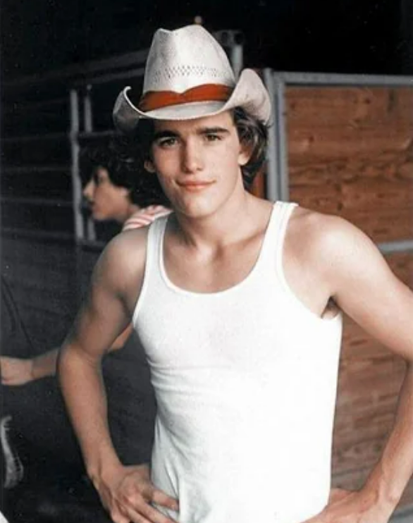 A young man wearing a white tank top and a cowboy hat stands with his hands on his hips, smiling slightly. There is a wooden wall and another person blurred in the background.