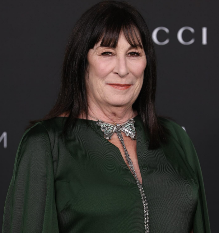 A woman with straight dark hair and bangs, wearing a dark green dress with a jeweled neckline, smiles at a formal event. The background is dark with partially visible white text.