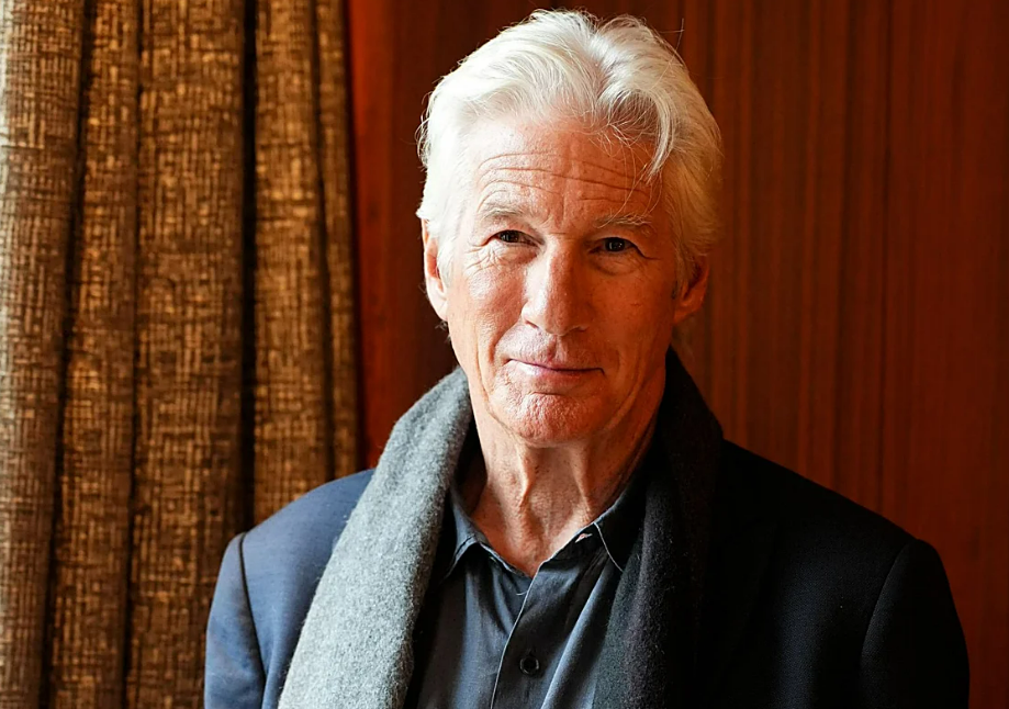 An older man with white hair and a slight smile stands indoors, wearing a dark jacket, a black shirt, and a gray scarf, with a wooden wall and brown curtain in the background.