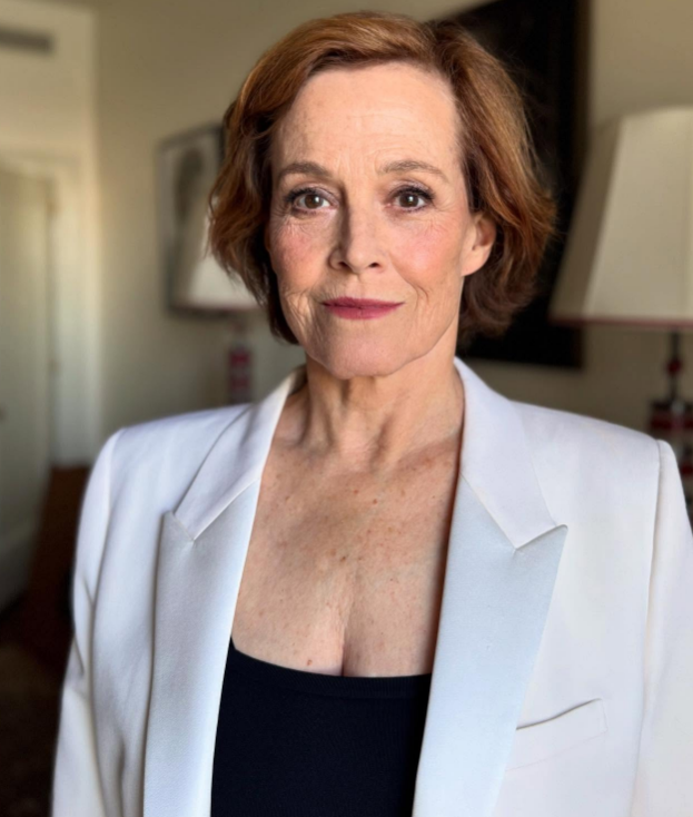 A woman with short, light brown hair wears a white blazer over a black top, standing indoors with a neutral expression. The background includes a door, a blurred painting, and two lamps.
