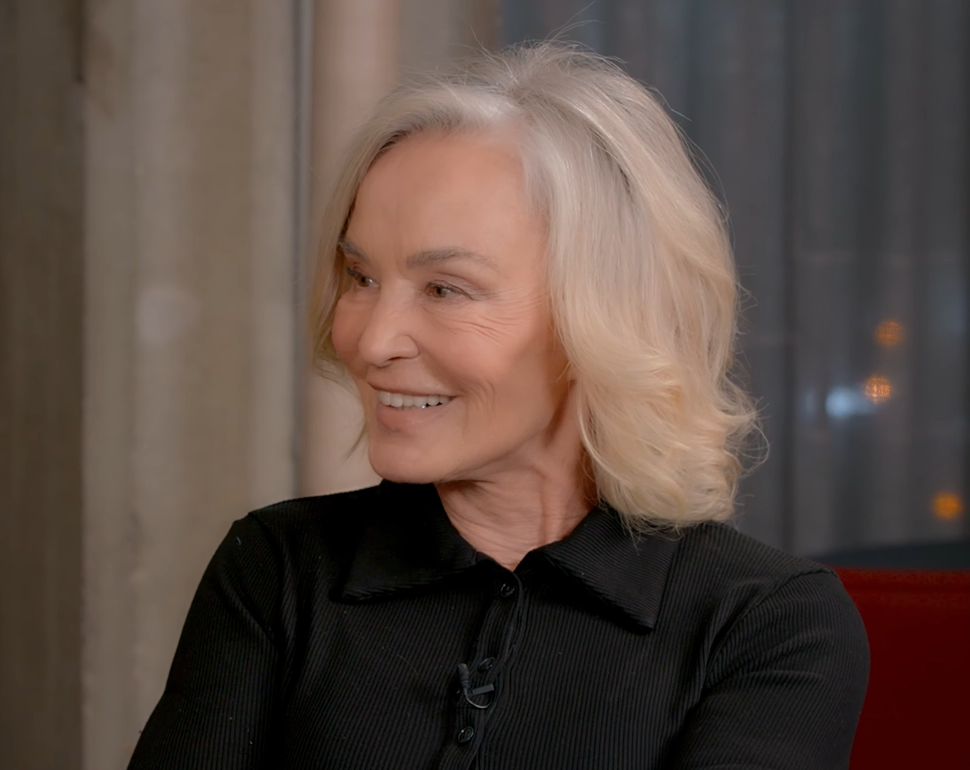 A smiling older woman with shoulder-length blonde hair wearing a black collared top sits indoors, facing slightly to the right.