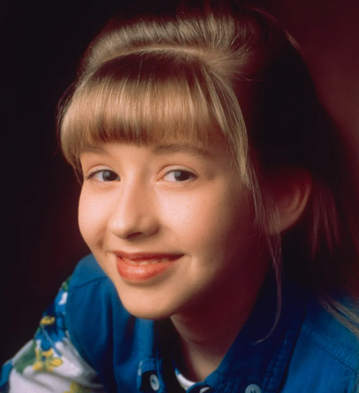 A young girl with straight blonde hair and bangs smiles softly. She is wearing a blue collared shirt over a patterned top, and the background is dark and out of focus.