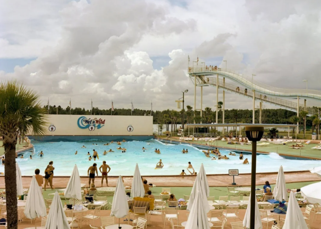 People swim and play in a large wave pool at a water park beneath a cloudy sky. The scene includes a tall water slide, palm trees, and rows of empty lounge chairs with white umbrellas in the foreground.