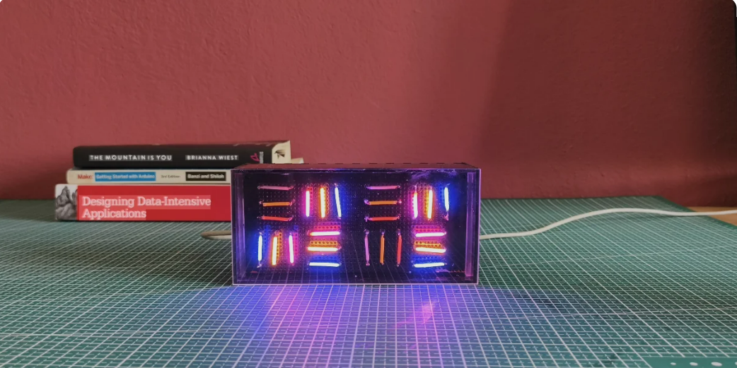 A rectangular digital clock with colorful LED lights displays the time on a grid cutting mat, with a stack of books and a red wall in the background. A white cable is attached to the clock.