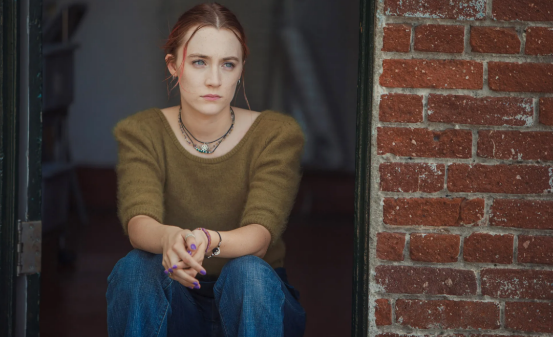 A young woman with light skin and auburn hair sits on a doorstep, wearing a green sweater and blue jeans. She looks serious and thoughtful, with her hands clasped in front of her. A brick wall is visible beside her.