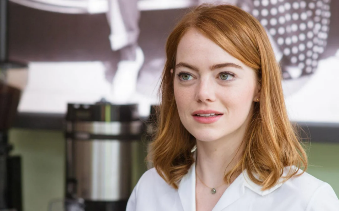 A woman with shoulder-length red hair and fair skin, wearing a white shirt and a delicate necklace, stands indoors with a blurred background including a coffee machine.