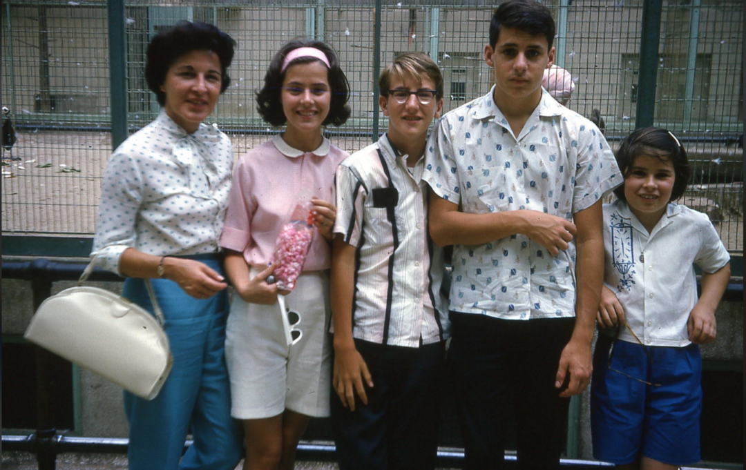 A woman and four children stand close together, smiling, near a fenced outdoor area. They are dressed in casual summer clothes. The woman holds a white purse; one girl holds a pink item. The background includes a metal fence and concrete.