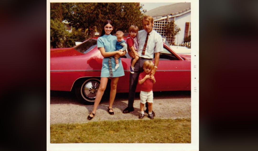 A young family of five stands in front of a red vintage car. The mother holds a baby, the father wears a tie, and two small children stand in front. Trees and a house are visible in the background.