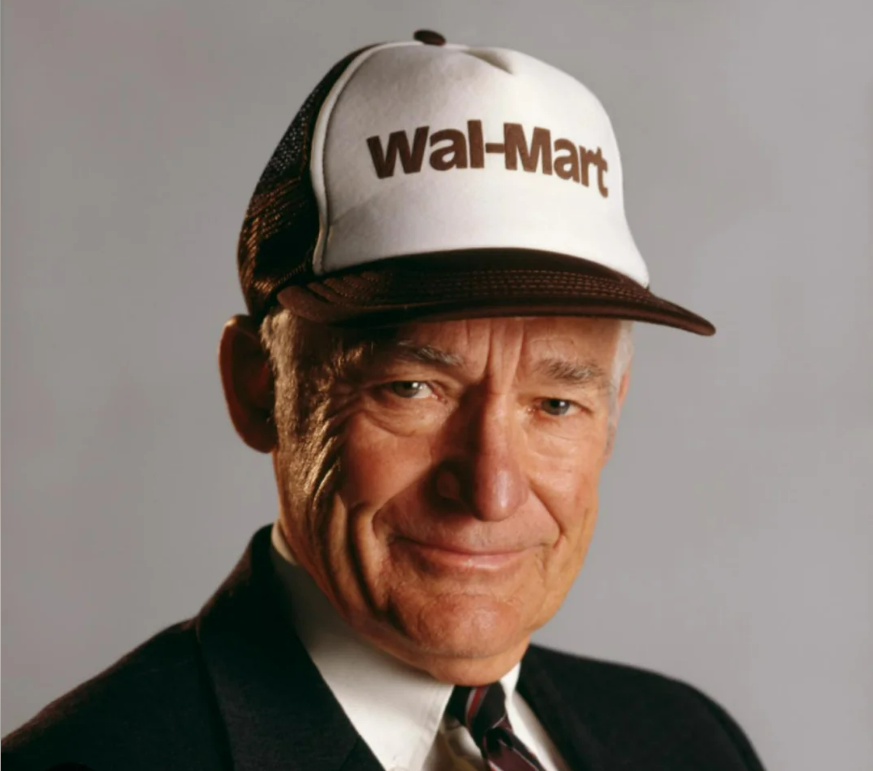 An older man wearing a suit, tie, and a white and brown cap with "Wal-Mart" written on it, smiling at the camera against a plain background.