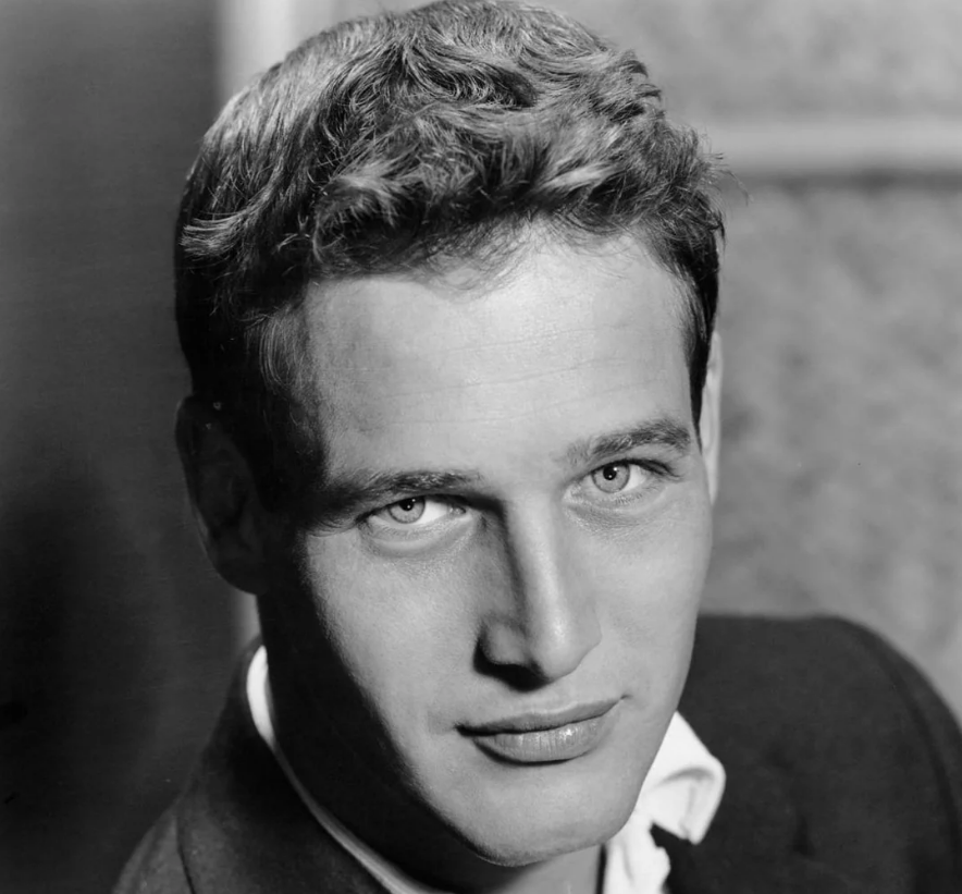 Black-and-white portrait of a young man with light eyes and wavy hair, wearing a dark jacket over a white collared shirt, looking directly at the camera with a slight smile.