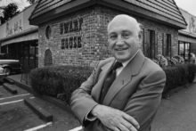 A smiling man in a suit stands with arms crossed in front of a Waffle House restaurant, with cars parked nearby and trees in the background.
