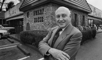A smiling man in a suit stands with arms crossed in front of a Waffle House restaurant, with cars parked nearby and trees in the background.