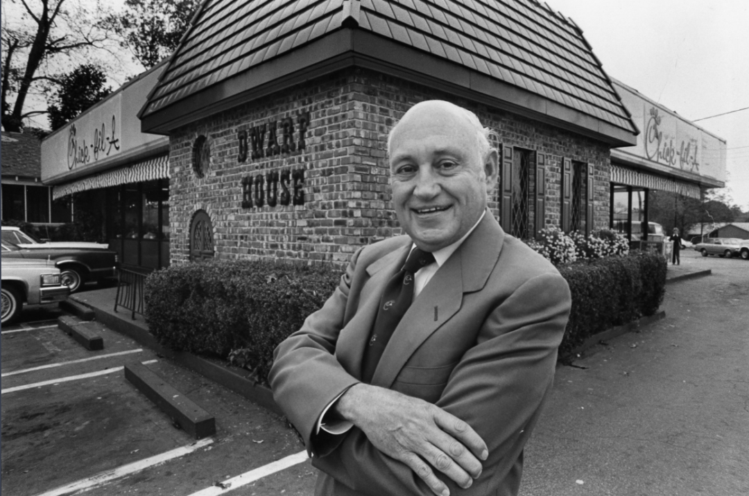 A smiling man in a suit stands with arms crossed in front of a brick Waffle House restaurant. Cars are parked nearby, and the restaurant sign is visible on the building. Black-and-white photo.