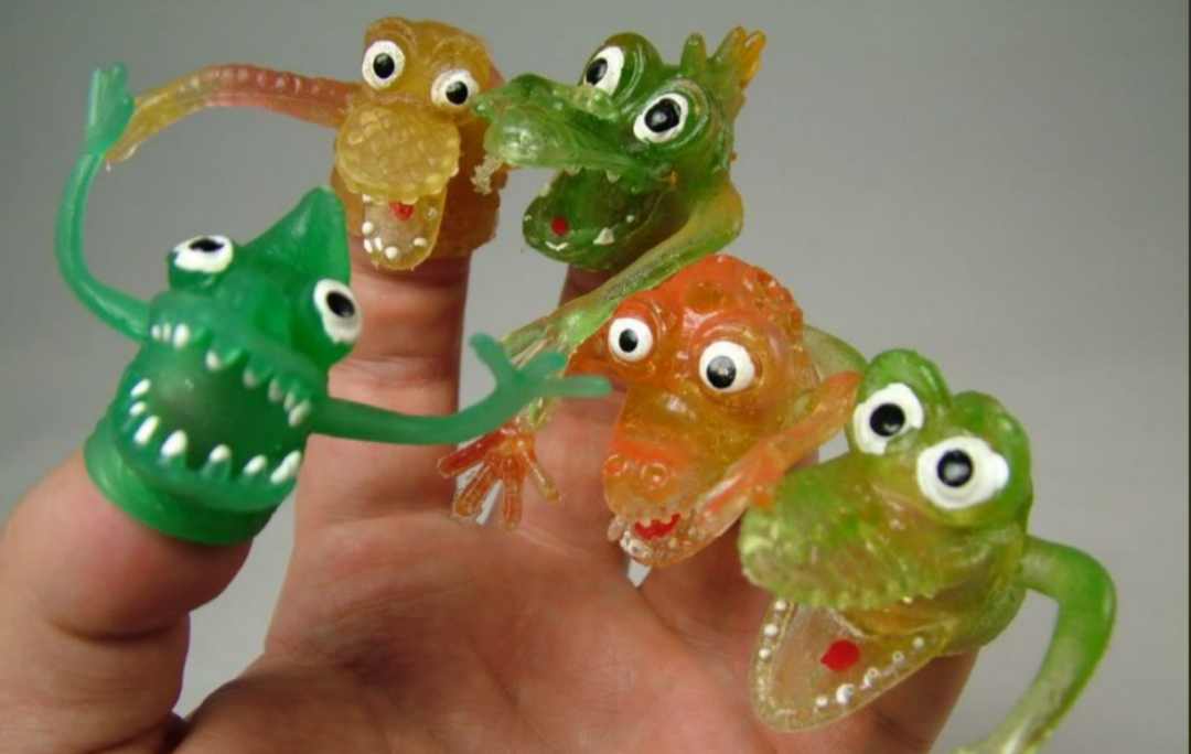 A hand with five fingers, each wearing a colorful, translucent, rubber monster finger puppet with big googly eyes and open mouths. The puppets are green, yellow, or orange.
