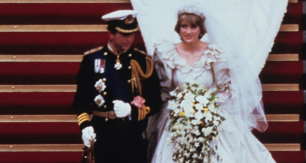 A bride in a white wedding gown with a long veil and large bouquet walks arm in arm with a groom in a decorated navy military uniform down red-carpeted steps.