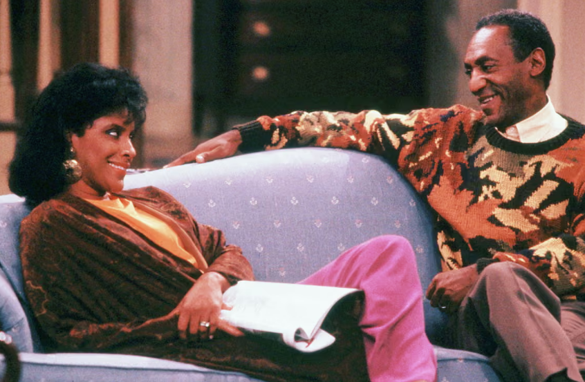 A woman and a man sit on a light blue sofa, smiling and talking. The woman is wearing a dark jacket and pink pants, holding papers, while the man wears a colorful patterned sweater.