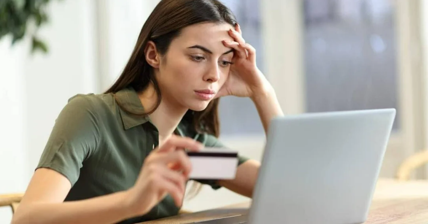 A woman sits at a table, looking concerned while holding a credit card and staring at a laptop screen, suggesting she may be worried about an online purchase or financial issue.