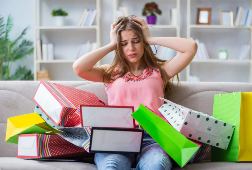 A young woman sits on a couch holding her head in frustration, surrounded by colorful shopping bags and boxes, with shelves and plants in the background.