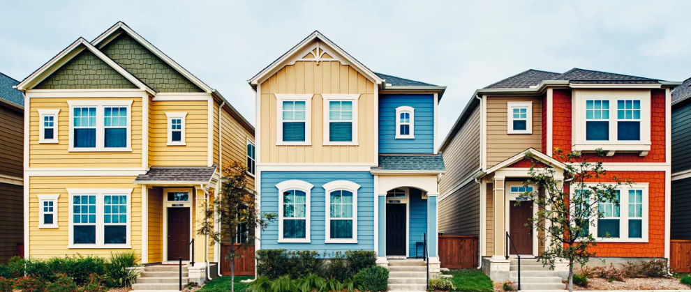 Three colorful, two-story suburban houses stand side by side. The left house is yellow with green accents, the middle house is blue with white trim, and the right house is orange with brown trim. Small trees line the front yards.