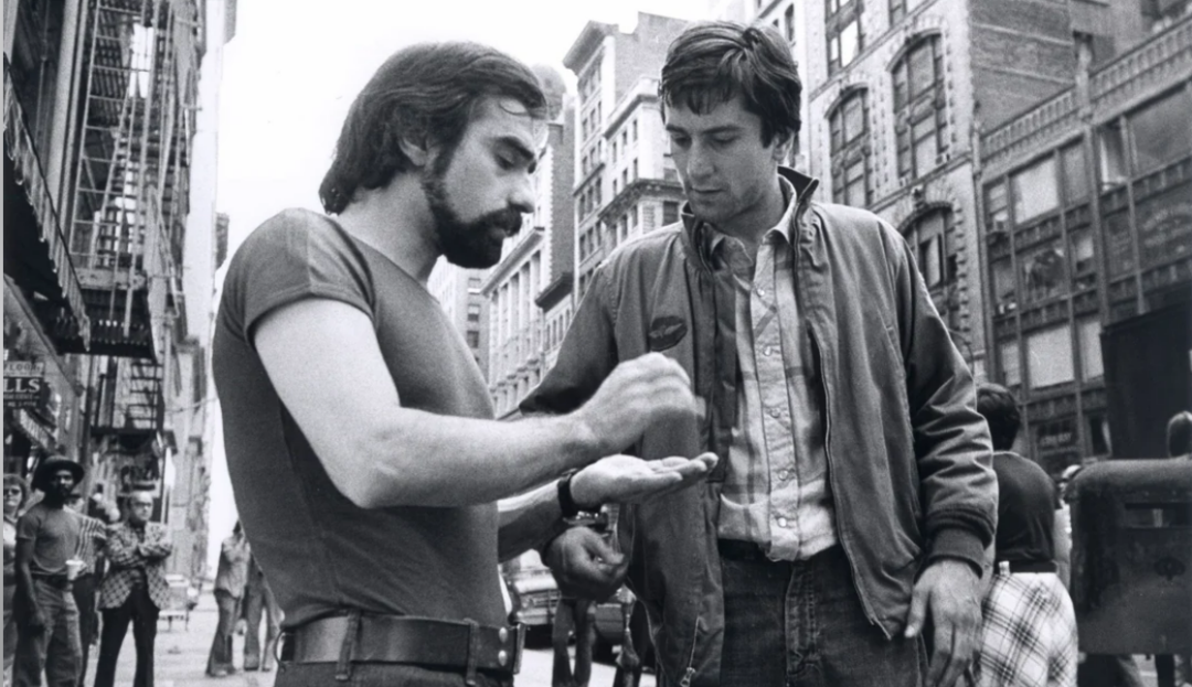 Two men stand on a busy city sidewalk; one examines something in his hand while the other watches. Tall buildings and pedestrians are visible in the background, creating an urban atmosphere.