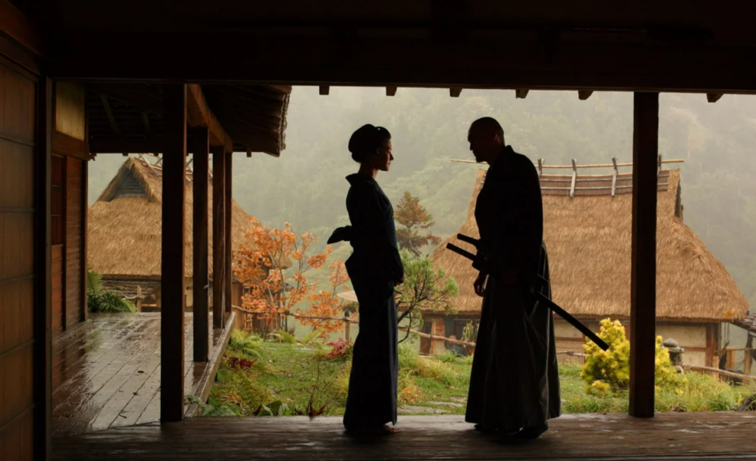 Two people in traditional Japanese attire stand facing each other on a wooden porch, with thatched-roof houses and autumn trees visible in the misty background.