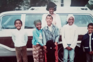 Six smiling children stand in front of a vintage station wagon, with one adult woman standing behind them. They are dressed in colorful, retro clothing, and a black dog is at the center of the group.