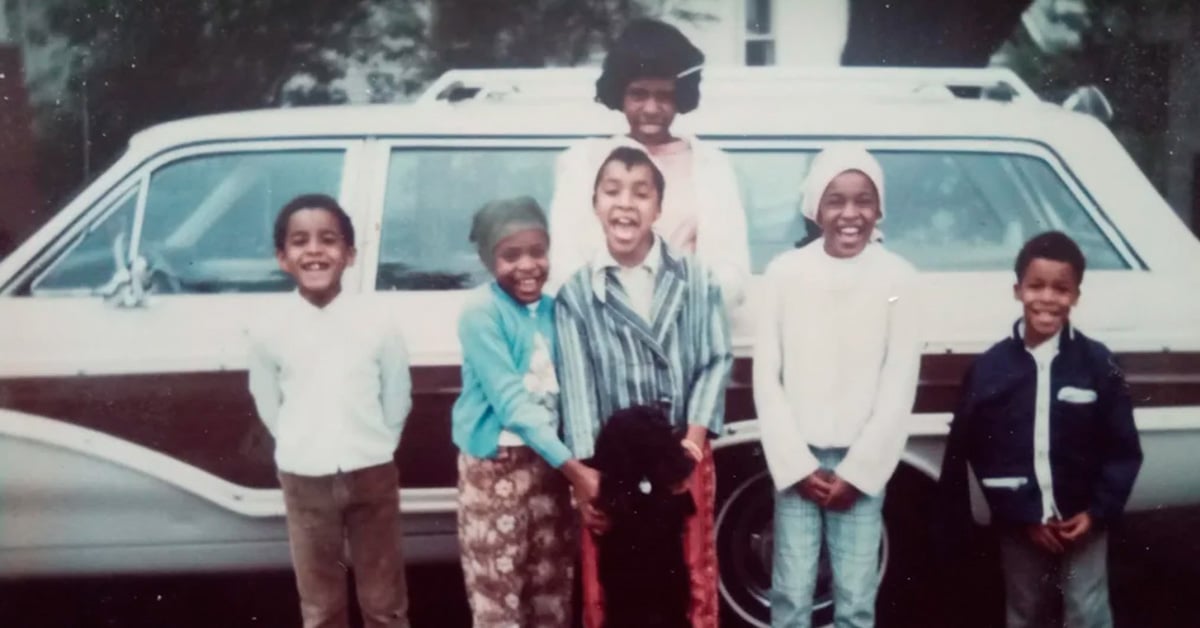 Six smiling children stand in front of a vintage station wagon, with one adult woman standing behind them. They are dressed in colorful, retro clothing, and a black dog is at the center of the group.