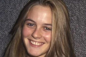 A young woman with straight, light brown hair smiles at the camera against a textured gray background. She is wearing a light-colored top.